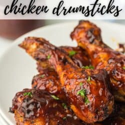 A close-up of juicy, glazed air fryer BBQ drumsticks on a white plate, garnished with herbs. A jar of BBQ sauce peeks from the background. Text on the image reads Air Fryer BBQ Chicken Drumsticks and HomemadeHeather.com.
