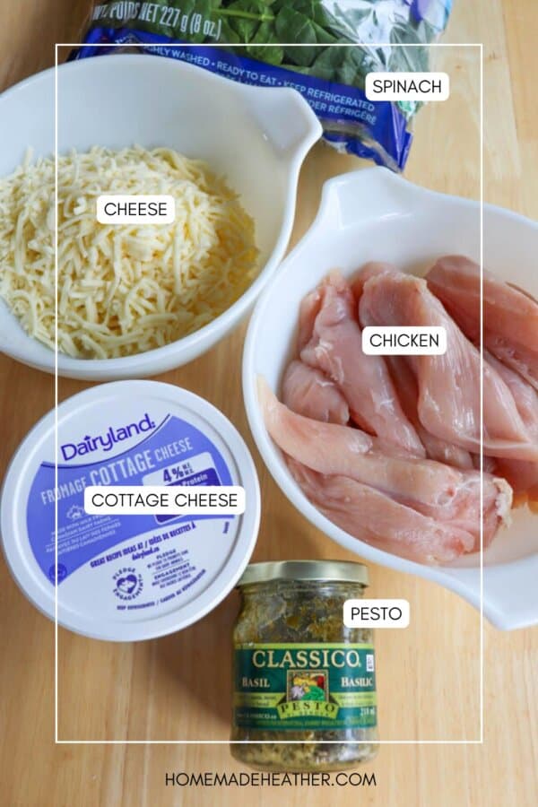 Ingredients on a wooden surface: a bag of spinach, bowls of shredded cheese and raw chicken strips, a container of cottage cheese, and a jar of basil pesto.