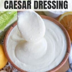 A bowl of creamy cottage cheese Caesar dressing with a wooden spoon lifting a portion. Surrounded by fresh lettuce, lemon halves, and garlic cloves. Text overlay reads cottage cheese Caesar dressing. Website URL at the bottom.