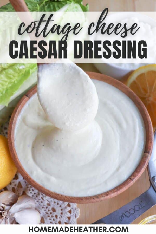 A bowl of creamy cottage cheese Caesar dressing with a wooden spoon lifting a portion. Surrounded by fresh lettuce, lemon halves, and garlic cloves. Text overlay reads cottage cheese Caesar dressing. Website URL at the bottom.