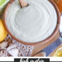 A wooden bowl of creamy Caesar dressing with a wooden spoon, surrounded by lemon, garlic cloves, lettuce, and a small bowl of grated cheese. Text reads high protein Caesar dressing and homemadeheather.com.