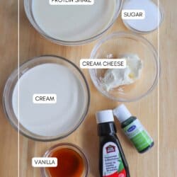 Top-down view of ingredients in bowls and bottles on a wooden surface. Visible items are labeled: protein shake, cream, cream cheese, sugar, vanilla, and mint extract. The text homemadeheather.com appears at the bottom.