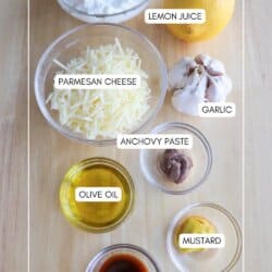 Ingredients for a recipe are arranged on a wooden surface: cottage cheese, a lemon, garlic, grated parmesan cheese, anchovy paste, olive oil, mustard, and Worcestershire sauce, each labeled with text.