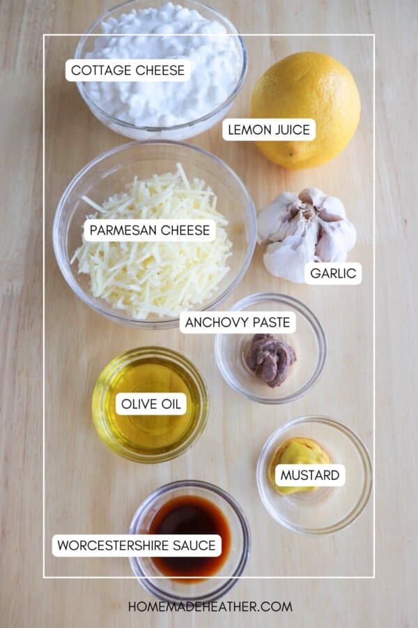 Ingredients for a recipe are arranged on a wooden surface: cottage cheese, a lemon, garlic, grated parmesan cheese, anchovy paste, olive oil, mustard, and Worcestershire sauce, each labeled with text.