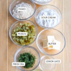 A wooden surface is topped with seven bowls containing various ingredients: cottage cheese, panko, sour cream, chopped pickles, butter cubes, fresh dill, and lemon juice, along with a label indicating pickle juice.