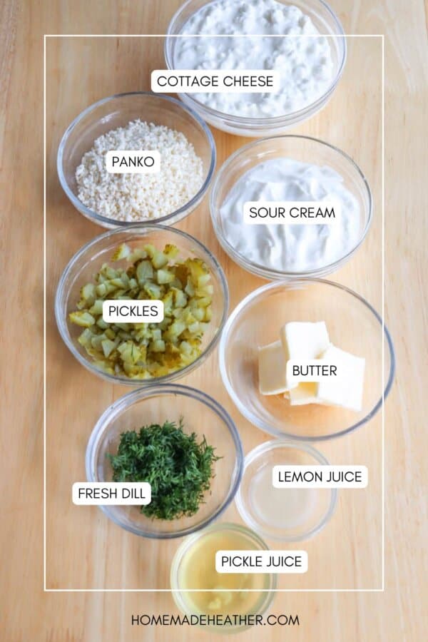 A wooden surface is topped with seven bowls containing various ingredients: cottage cheese, panko, sour cream, chopped pickles, butter cubes, fresh dill, and lemon juice, along with a label indicating pickle juice.