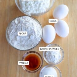 A wooden table with ingredients for a recipe: a bowl of cottage cheese, three eggs, a bowl of flour, a bowl of sugar, a bowl of baking powder, and a small bowl of vanilla extract. Each item has a label.