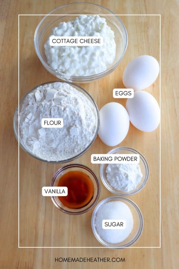 A wooden table with ingredients for a recipe: a bowl of cottage cheese, three eggs, a bowl of flour, a bowl of sugar, a bowl of baking powder, and a small bowl of vanilla extract. Each item has a label.