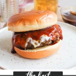 A juicy hamburger with BBQ sauce on a bun is on a white plate. A glass of beer and a sauce bowl are blurred in the background. The text at the bottom reads The Best BBQ Hamburger and homemadeheather.com.