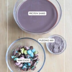 Bowls on a wooden table with labeled contents: a large bowl of brown liquid marked protein shake, a small bowl filled with brown powder labeled pudding mix, and a medium bowl packed with colorful candies labeled bunny tracks mini eggs.