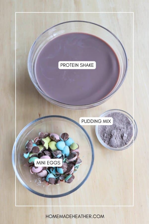 Bowls on a wooden table with labeled contents: a large bowl of brown liquid marked protein shake, a small bowl filled with brown powder labeled pudding mix, and a medium bowl packed with colorful candies labeled bunny tracks mini eggs.