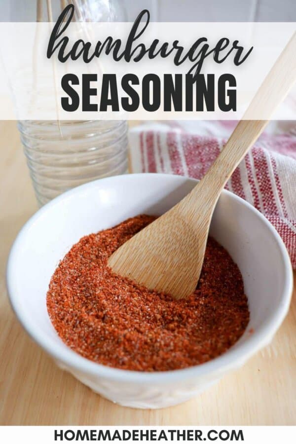 A bowl of reddish-brown hamburger seasoning with a wooden spoon resting in it, placed on a wooden surface. A striped cloth is partially visible in the background. Text on the image reads hamburger seasoning and homemadeheather.com.