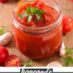 A jar of homemade salsa topped with parsley on a wooden surface. Surrounding the jar are cherry tomatoes, garlic cloves, and a piece of red and white checkered cloth. Text at the bottom reads homemade salsa canning homemadeheather.com.