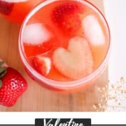 Close-up of two glasses filled with a red punch, ice, and strawberry slices. One slice resembles a heart. Fresh strawberries are scattered around. Text at the bottom reads: Valentine Punch Recipe homemadeheather.com.