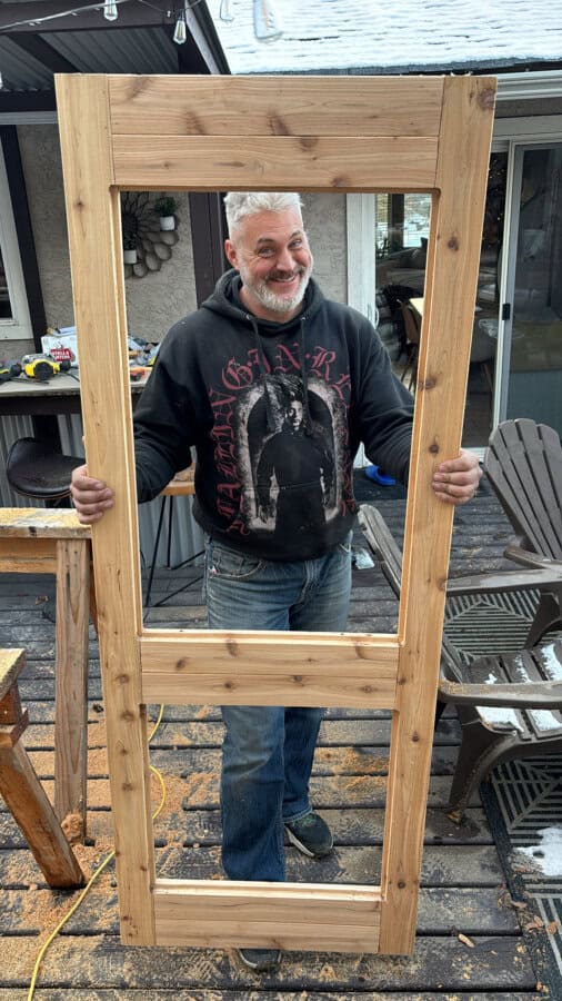 A person wearing a black hoodie with a graphic design holds a wooden door frame while smiling. The scene is outdoors, with woodworking tools and materials on a deck in the background.