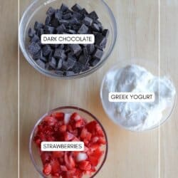 Three bowls on a wooden surface: one with dark chocolate chunks, one with Greek yogurt, and one with sliced strawberries. Labels on each bowl identify the contents.