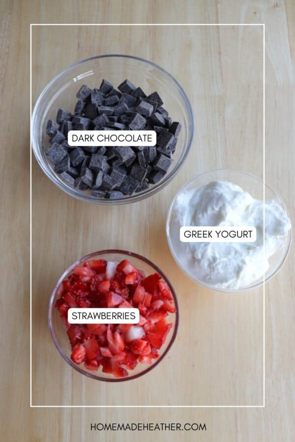 Three bowls on a wooden surface: one with dark chocolate chunks, one with Greek yogurt, and one with sliced strawberries. Labels on each bowl identify the contents.