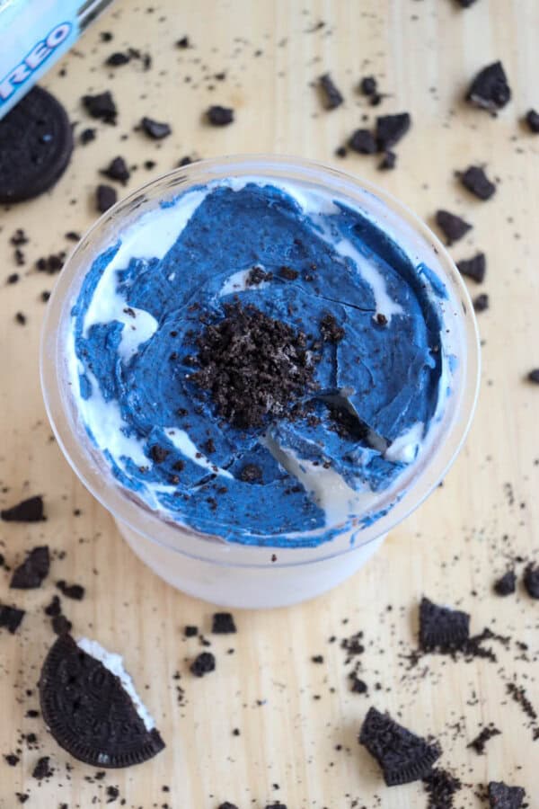 A cup of dessert with a swirl of blue and white cream topped with crumbled chocolate cookie pieces. Broken chocolate cookies are scattered on the wooden surface around the cup.
