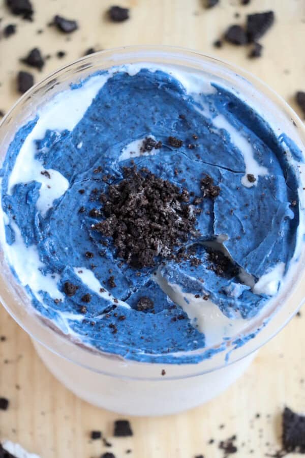 A pint of ice cream featuring layers of white cream and vibrant blue chocolate, topped with crumbled chocolate cookie bits. Cookie crumbs are scattered on a light wooden surface around the glass.