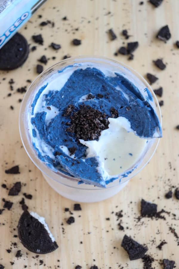 A cup of blue and white ice cream topped with crushed cookies, placed on a wooden surface. Broken pieces of chocolate sandwich cookies are scattered around, and a partially visible cookie package is in the corner.