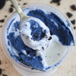 A top view of a spoon lifting a creamy dessert with a mix of blue and white colors from a cup. Crumbled cookies are scattered around on a light wooden surface.