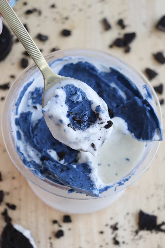 A top view of a spoon lifting a creamy dessert with a mix of blue and white colors from a cup. Crumbled cookies are scattered around on a light wooden surface.