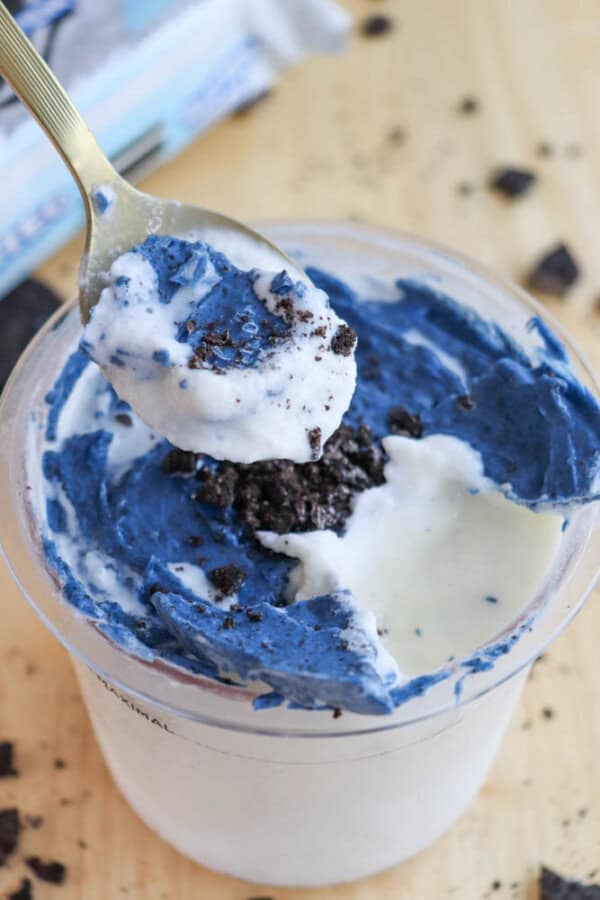 A spoonful of vanilla and blue ice cream is being lifted from a cup. The ice cream is mixed with crumbled cookies, with a creamy texture and cookie pieces visible throughout. The cup is placed on a light wooden surface.