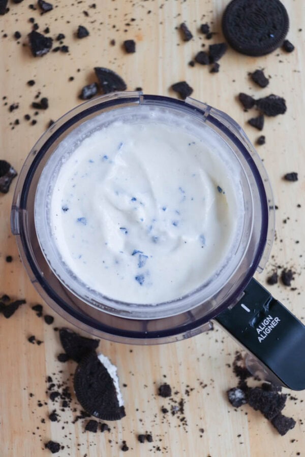 A food processor blending creamy white ice cream with visible cookie pieces. Broken chocolate cookies are scattered around the processor on a wooden surface.