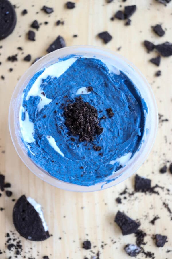 A container of soft serve-like ice cream topped with blue chocolate and cookie crumbs sits on a wooden surface. Crumbled chocolate cookies are scattered around.
