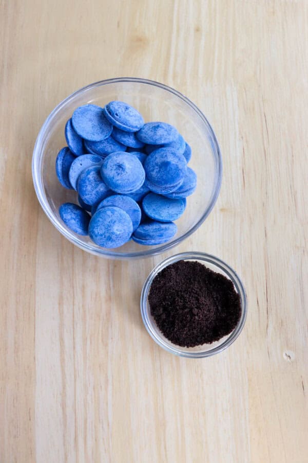 A glass bowl filled with blue chocolate wafers is placed next to a smaller glass bowl containing ground coffee on a light wooden surface.