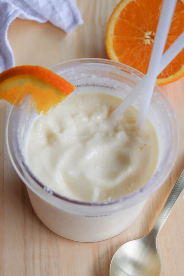 A creamy orange smoothie in a clear cup is garnished with a slice of orange. Two white straws are in the drink. A silver spoon and a halved orange are beside the cup on a light wooden surface.