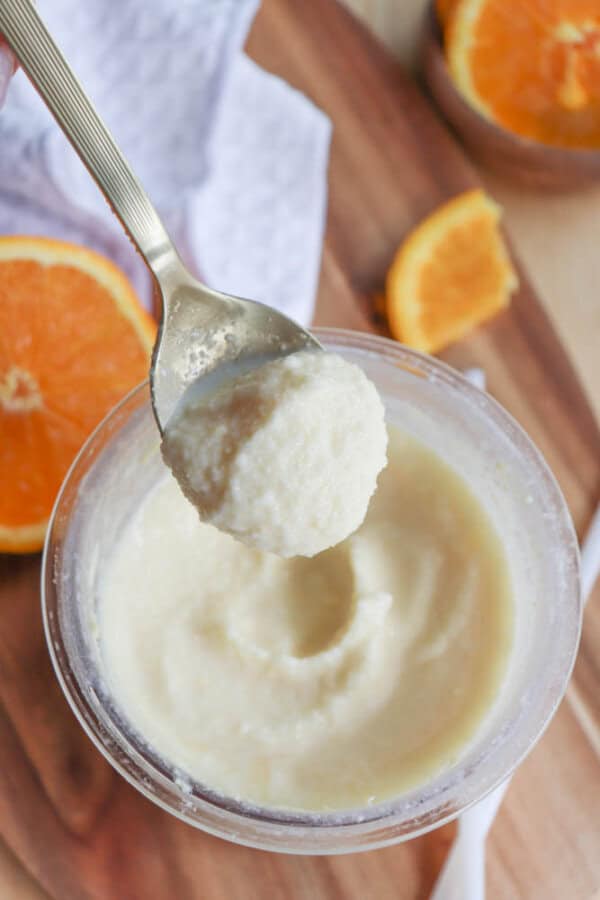 A spoonful of creamy, light-colored sorbet is held above a clear container filled with the same dessert. Fresh orange slices and a white napkin are nearby on a wooden surface.