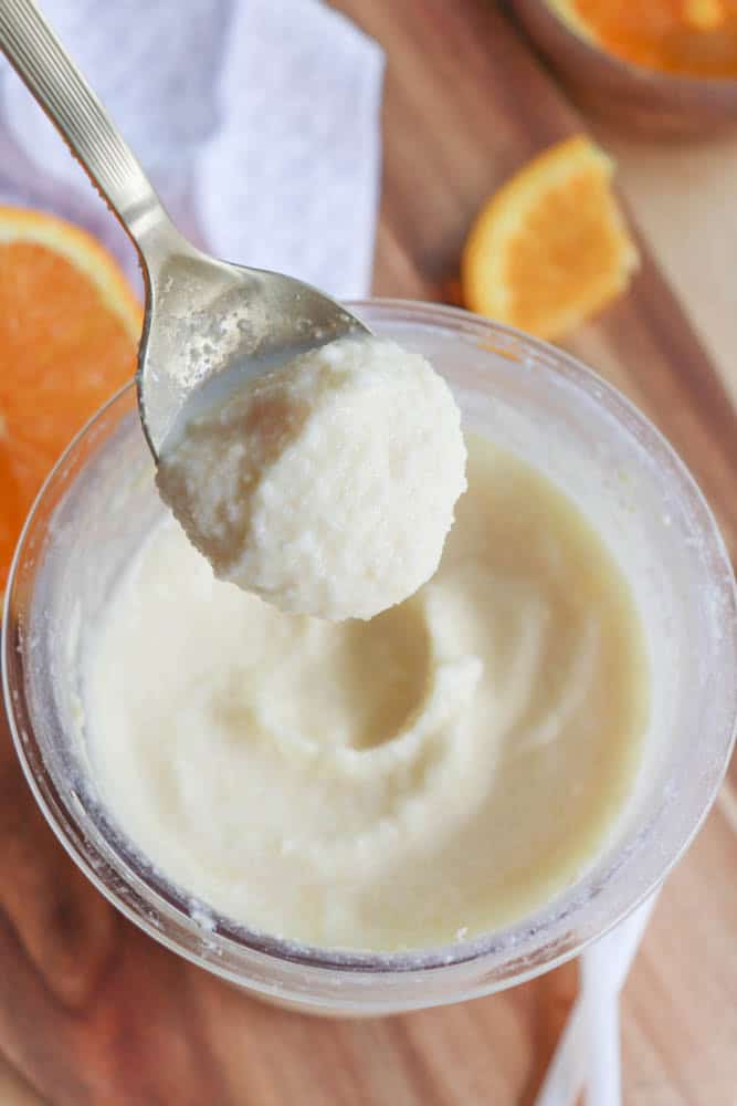 A spoon scooping out a creamy, white mixture from a clear jar placed on a wooden surface. Orange slices and a white cloth are partially visible in the background.