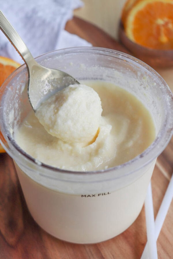 A spoon scooping creamy, light-colored sorbet from a clear container. The container is filled close to the max fill line. In the background, there are slices of an orange and a white cloth on a wooden surface.