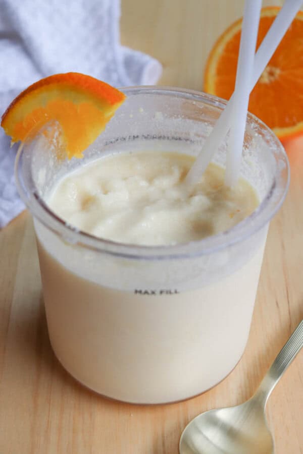 A creamy orange smoothie is in a clear plastic cup with two white straws. An orange slice garnishes the rim. A spoon and a half orange are nearby on a wooden surface. A textured white cloth is in the background.