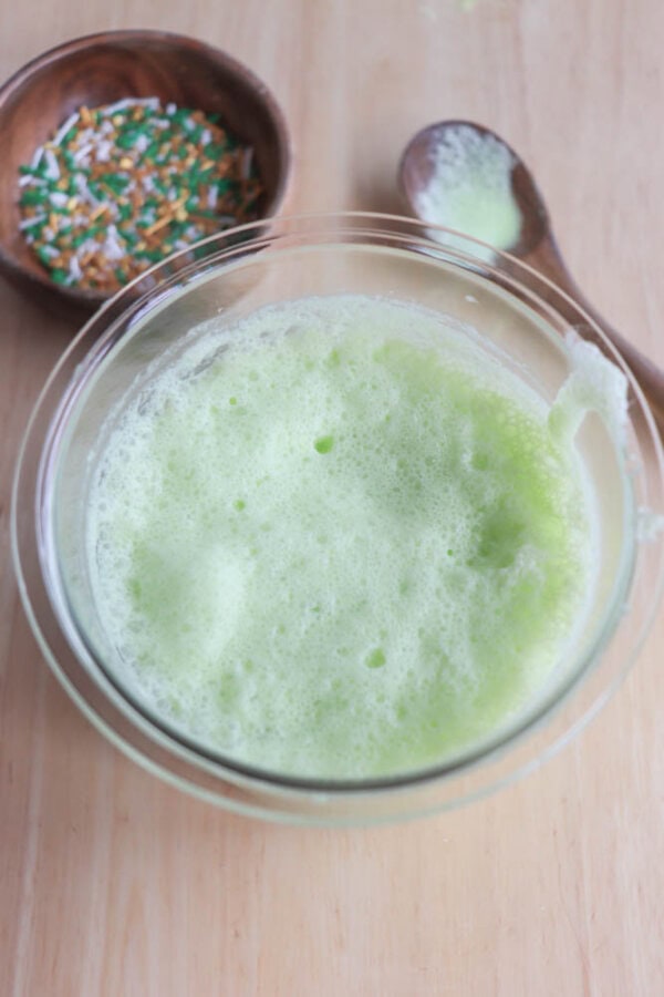 A glass bowl filled with frothy, light green foam sits on a wooden table. Nearby, a wooden spoon and a small bowl contain colorful sprinkles, predominantly green, orange, and white.