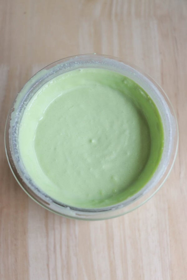 A clear glass bowl filled with smooth, light green pudding resembling a shamrock shake is placed on a wooden surface. The creamy texture of this delightful creation occupies nearly the entire bowl.