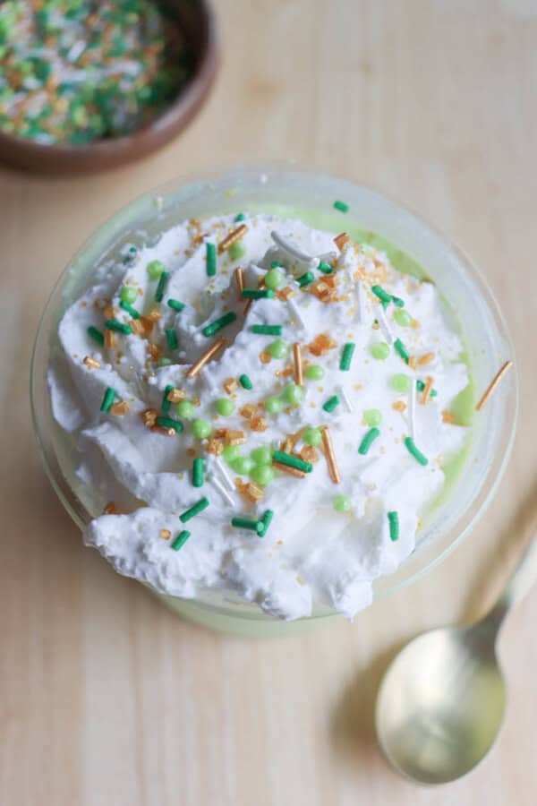 A shamrock shake dessert topped with whipped cream and green, gold, and white sprinkles sits elegantly in a glass on a wooden surface. Beside it lies a gold spoon, ready for action. In the background, a small bowl of sprinkles stands by like a ninja recipe secret weapon.