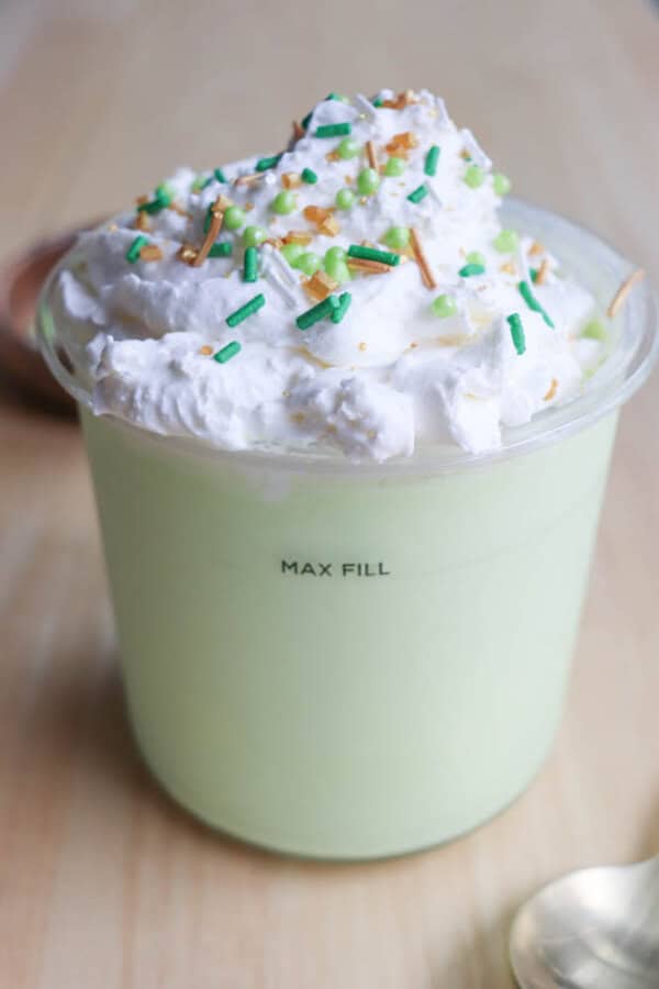 A plastic cup filled with light green pudding resembling a shamrock shake, topped with whipped cream and colorful sprinkles. The cup is marked Max Fill, hinting at a ninja recipe. A spoon lies beside it on a wooden surface.