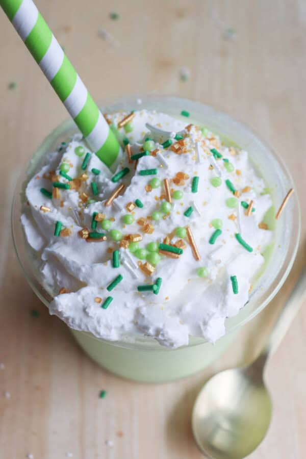 A creamy beverage, like a ninja-crafted Shamrock Shake, is topped with whipped cream and green, gold, and white sprinkles. A green and white striped straw is inserted into the drink in a clear cup. A gold spoon lies beside it on a light wooden surface.