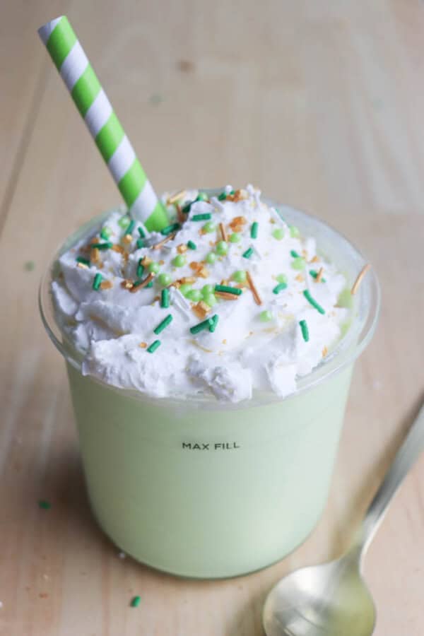 A creamy shamrock shake topped with whipped cream, green and yellow sprinkles. Adorned with a green-striped straw, it sits on a wooden surface beside a spoon. The word MAX FILL is visible on the cup—a true ninja recipe delight.
