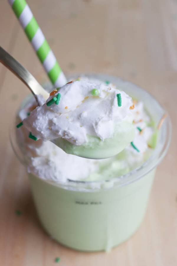 A spoonful of creamy, pale green shamrock shake topped with whipped cream and colorful sprinkles is held above a cup. The cup, also filled with the milkshake, has a green and white striped straw. The surface beneath is light in color.