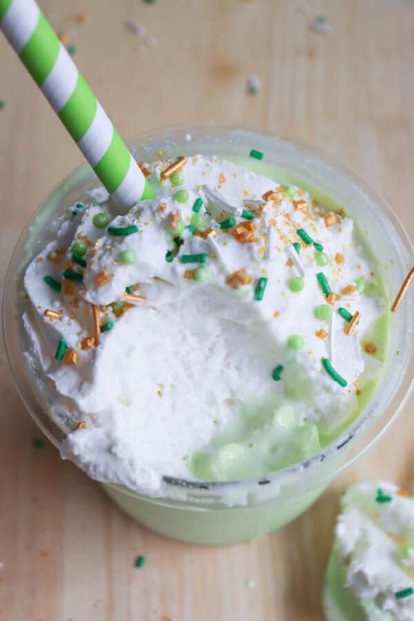 A creamy shamrock shake topped with whipped cream, green and gold sprinkles, and a green and white striped straw. The background is a wooden surface.