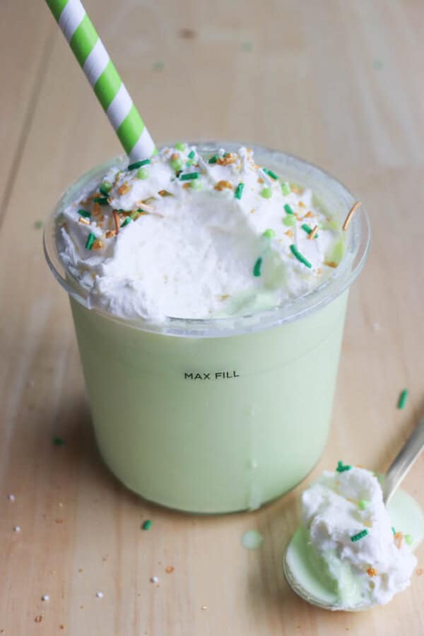 A light green shamrock shake topped with whipped cream, caramel drizzle, and green sprinkles in a clear plastic cup with a green and white striped straw. A spoon with whipped cream rests beside the cup on a wooden surface, showcasing this ninja recipe delight.