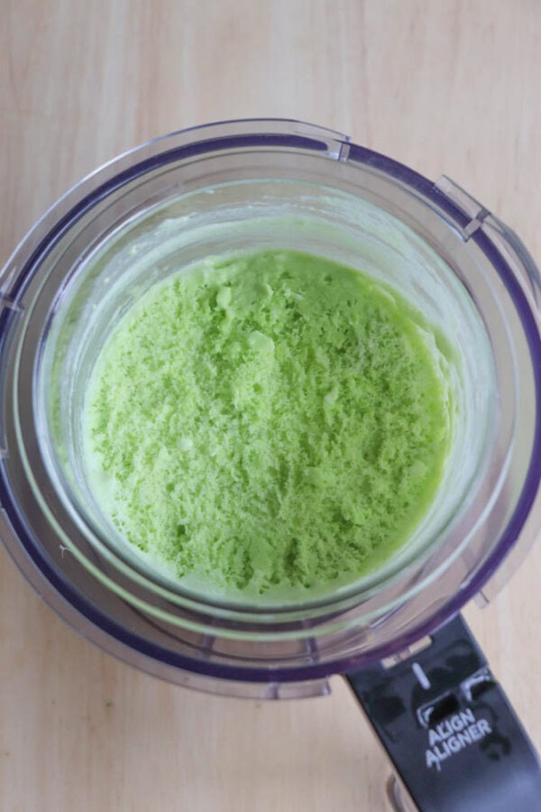 Top view of a ninja creami cup filled with bright green, creamy mixture, of a shake base, set on a light wooden surface. The mixture looks fluffy and smooth.