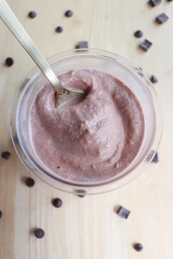 A glass cup filled with thick, creamy chocolate smoothie. A spoon is dipped into it. Chocolate chips are scattered around the cup on a light wooden surface.