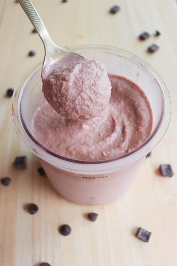 A spoon is lifting a scoop of light brown creamy dessert from a transparent cup. The cup is sitting on a light wooden surface, surrounded by scattered chocolate chips.