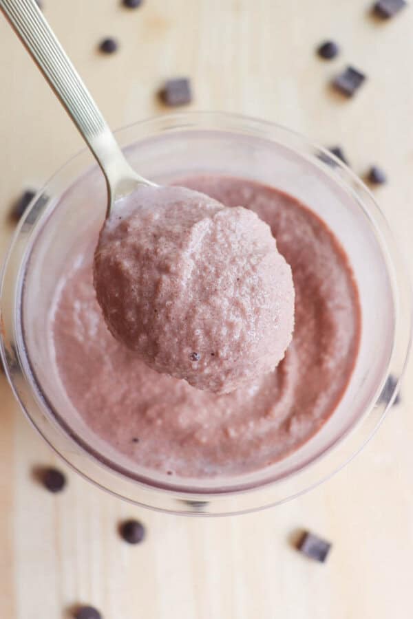 A spoonful of creamy chocolate mousse is held above a glass bowl filled with more mousse. Chocolate chips are scattered on a light wooden surface surrounding the bowl.