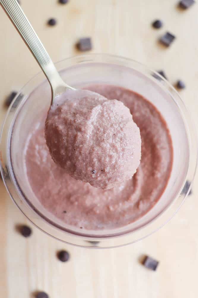 A spoonful of creamy chocolate mousse is held above a glass bowl filled with more mousse. Chocolate chips are scattered on a light wooden surface surrounding the bowl.