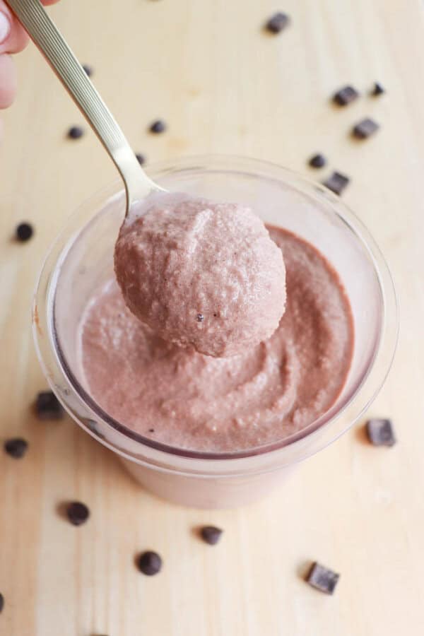 A spoon scooping chocolate mousse from a clear cup on a wooden surface. Chocolate chunks are scattered around the cup.